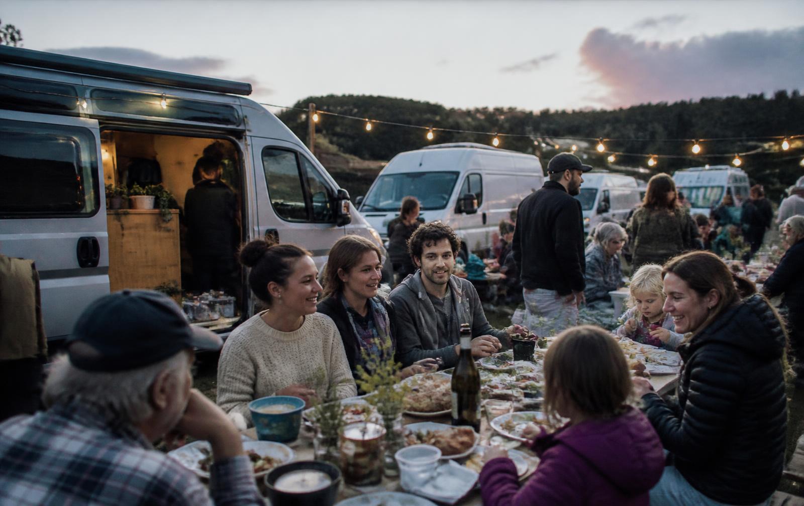 People of different ages sharing a long table beside camper vans at dusk